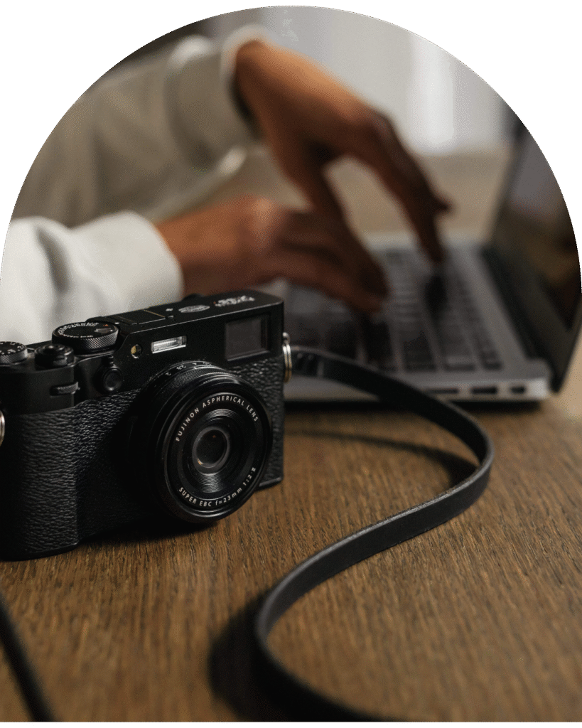 Na zdjęciu znajduje się aparat, w tle rozmazane dłonie piszące na klawiaturze laptopa